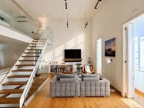 Ein modernes Wohnzimmer mit einem grauen Ecksofa, einer Treppe mit Glasgeländern, einem an der Wand montierten Flachbildfernseher und einem großen Fenster, das für natürliches Licht sorgt.