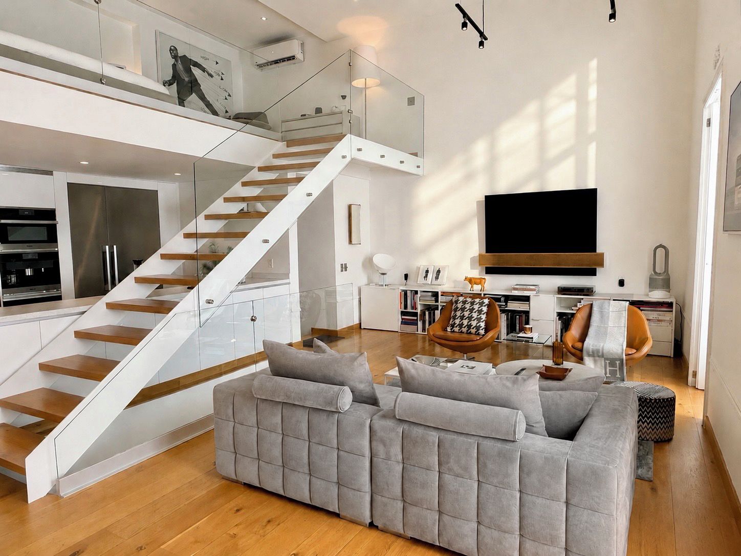 A modern living space with a staircase, a grey sofa, a flat-screen TV, and a kitchen area.
