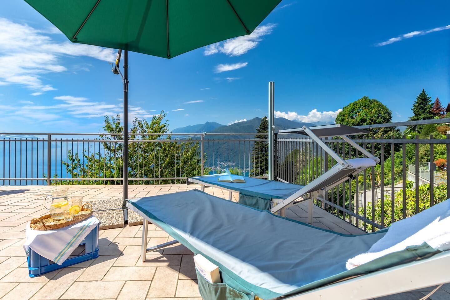 A balcony with a view of the sea and mountains, a table with a basket and a glass of water, and a lounge chair.