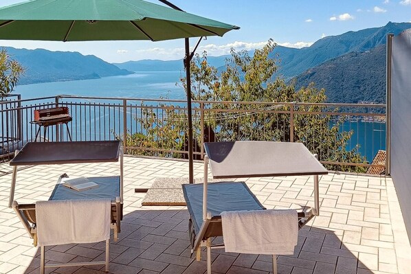 A terrace with two sun loungers, a barbecue grill, and a view of a lake and mountains. - Sunset View lago Maggiore (Maccagno Con Pino E Veddasca)