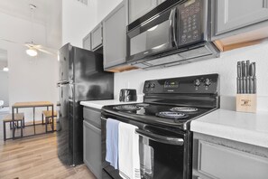 Een moderne keuken met een zwarte koelkast, een RVS magnetron en een zwart fornuis. Er staat een houten tafel met stoelen en er hangt een plafondventilator.
