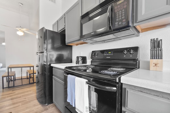 A modern kitchen with a black refrigerator, stainless steel microwave, and a black stove. There is a wooden table with chairs and a ceiling fan.