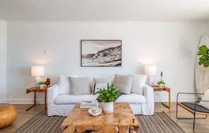 A modern living room with a white sofa, a coffee table with a plant, and a surfboard against the wall.