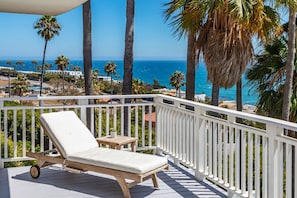 A deck with a lounge chair and a small table, overlooking a coastal area with palm trees and a view of the ocean.