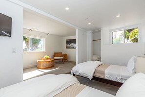 A hotel room with two beds, a TV, a window with a view of greenery, and a small seating area with a basket and chair.