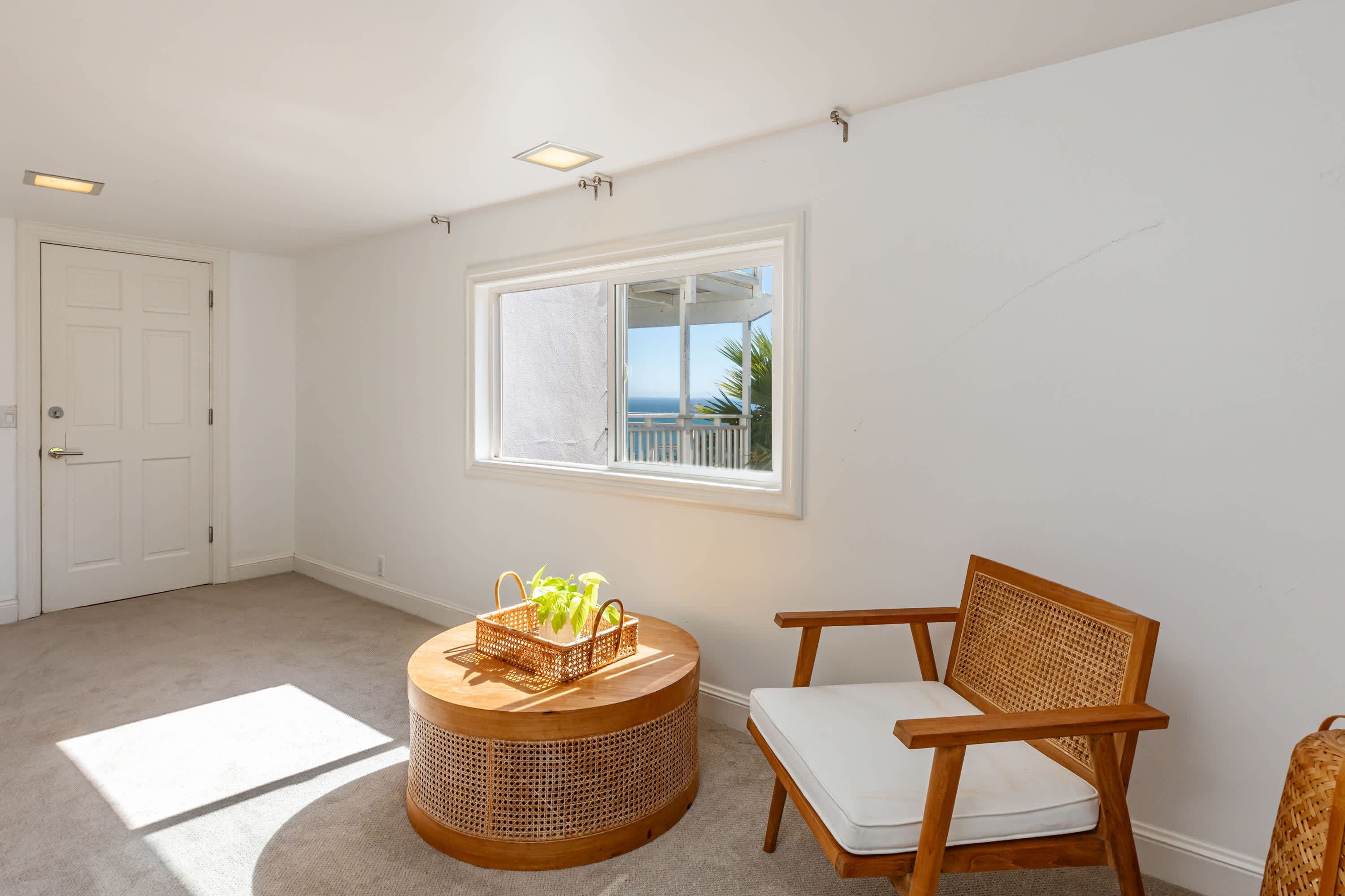 A room with a large window, a wicker basket with plants, a wooden chair, and a white door.