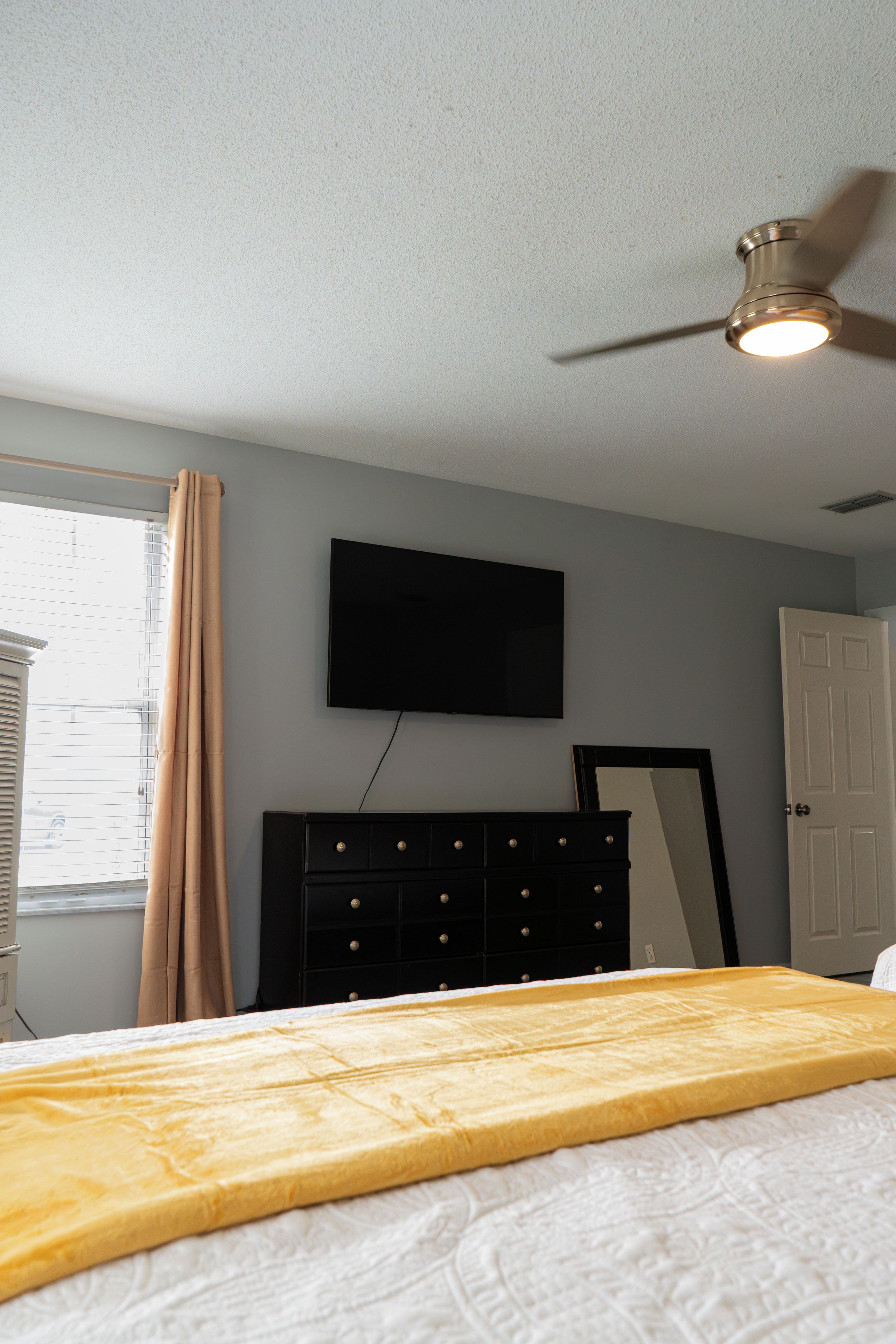A bedroom with a bed, a dresser, a television mounted on the wall, and a ceiling fan.