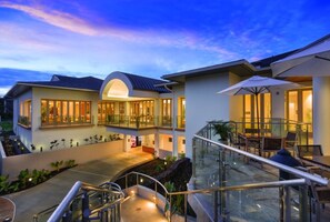Un hotel moderno con balaustrada de vidrio, mobiliario exterior y zona de piscina.