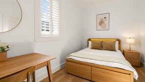 A bedroom with a wooden bed, bedside tables, a desk, and a window with blinds. - The Short Story Randwick - Unit 1 (Randwick)