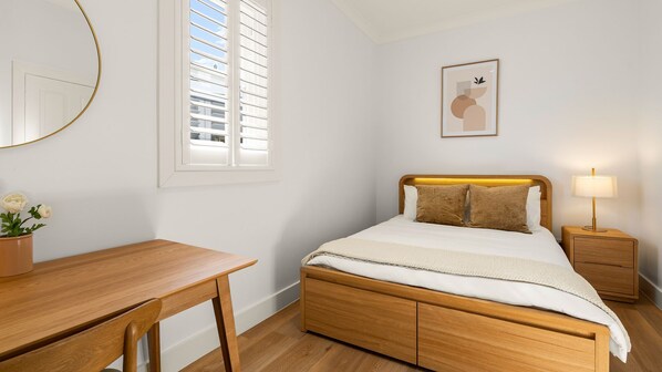 Ein Schlafzimmer mit einem Holzbett, Nachttischen, einem Schreibtisch und einem Fenster mit Jalousien.