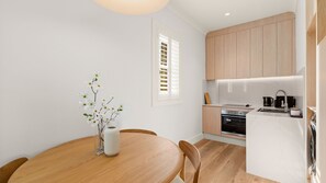 A compact kitchen with wooden cabinets, a dining table with chairs, and a vase with flowers. - The Short Story Randwick - Unit 1 (Randwick)