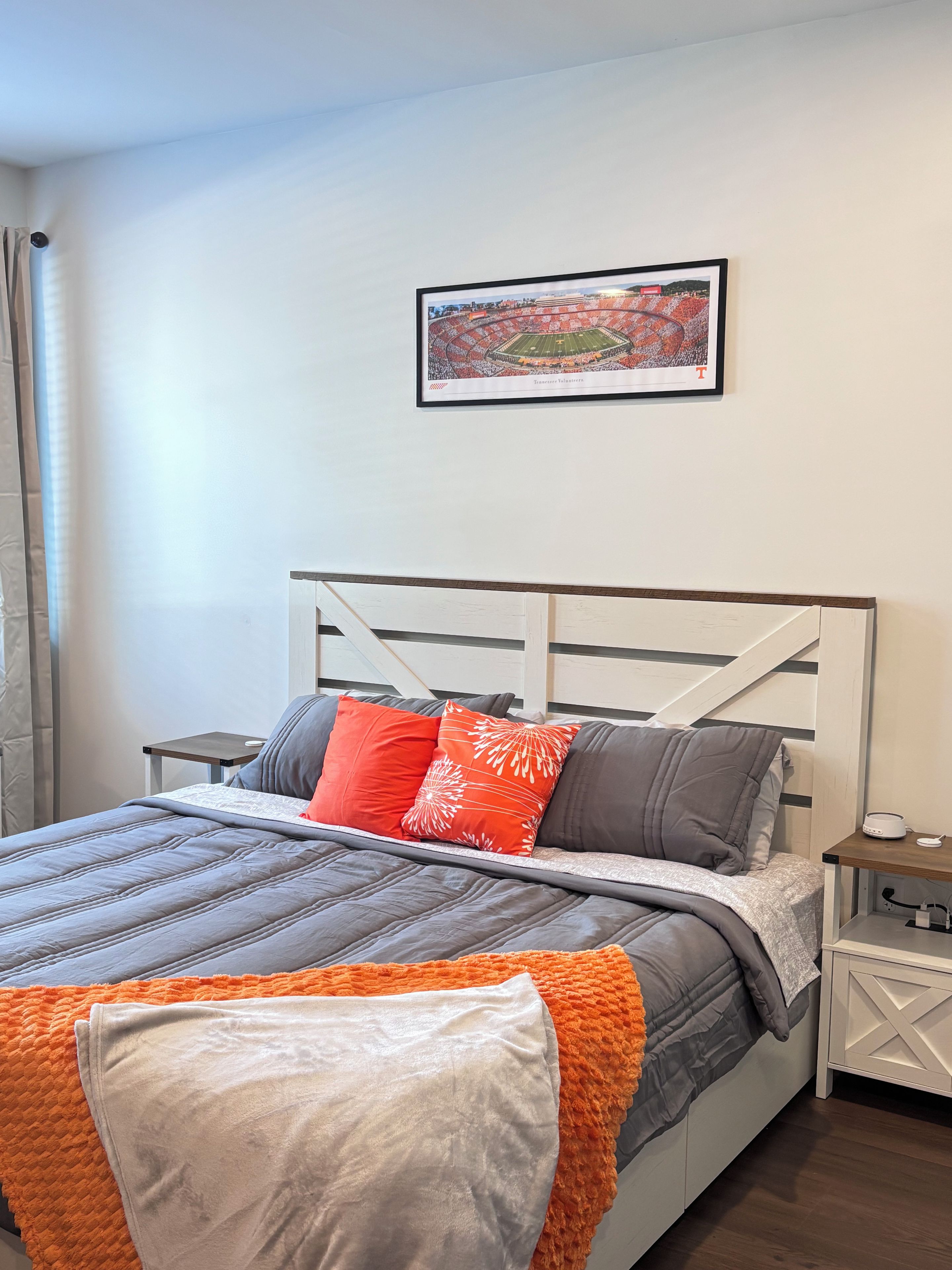 A bedroom with a bed, pillows, a bedside table, a nightstand, a framed picture on the wall, and a window with curtains.