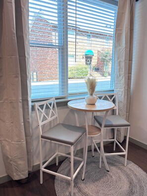A room with a round table and two chairs, a window with blinds, and a view of buildings outside.