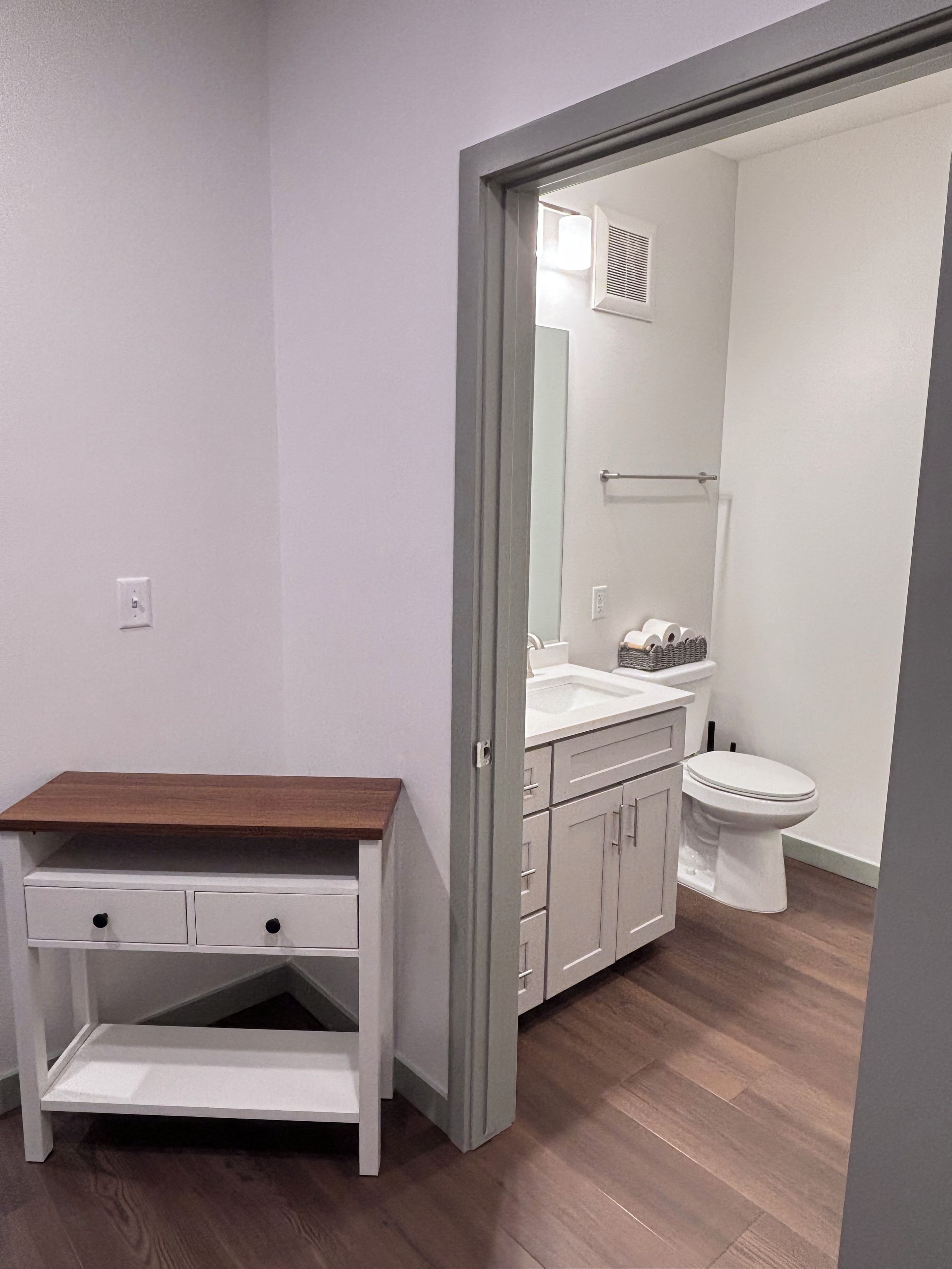 A bathroom with a toilet, sink, and mirror. There is a wooden shelf and a cabinet with drawers.