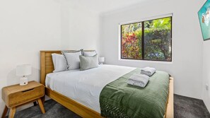 A bedroom with a wooden bed, white bedding, a green throw, a nightstand with a lamp, and a window with a view of foliage. - Surf's Up Sawtell - Walking Distance To Beach and Cafes (Sawtell)