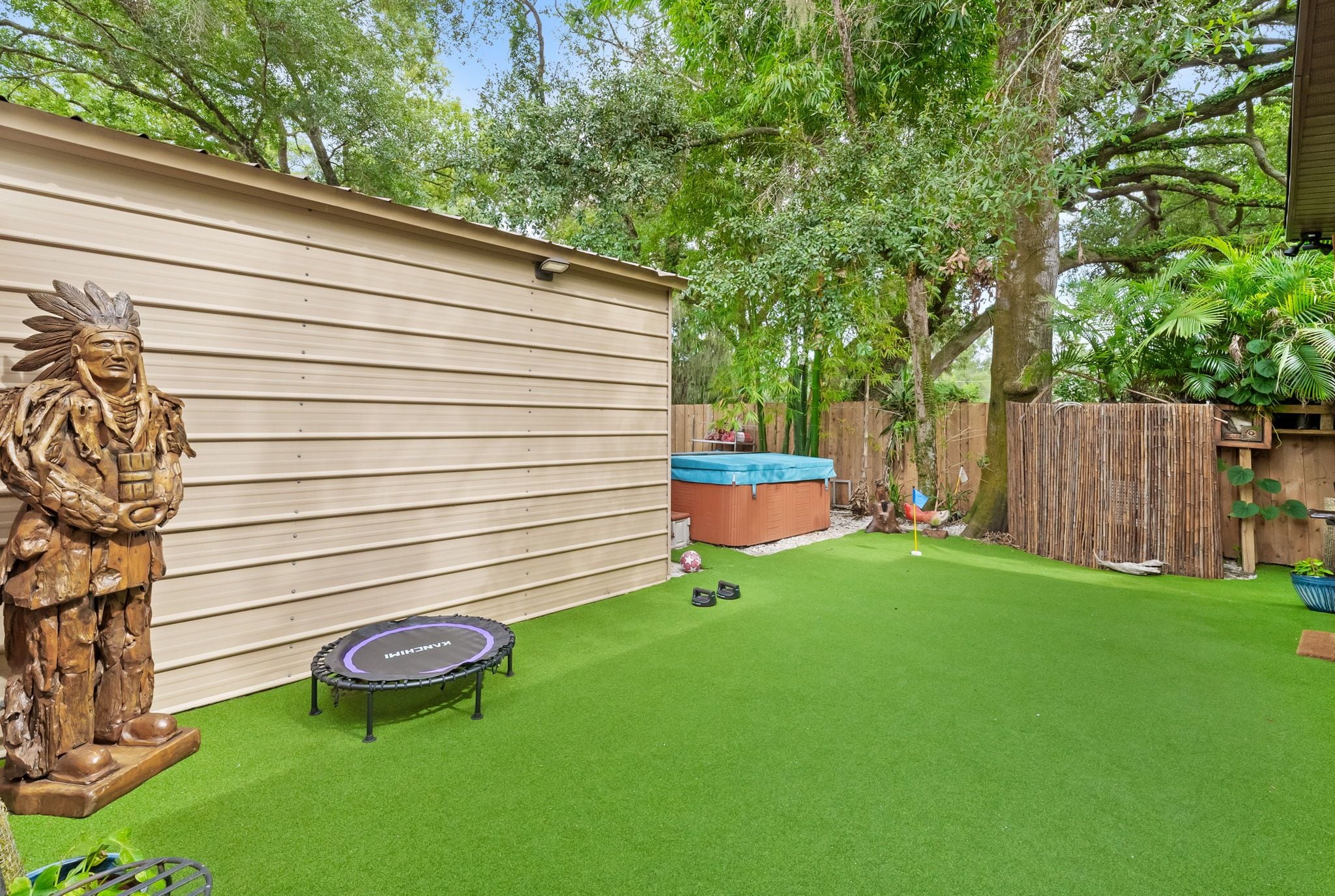Un patio trasero con césped artificial, un trampolín, un jacuzzi y una valla de madera.