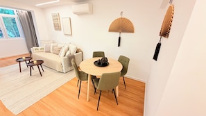 Ein modernes Wohnzimmer mit einer Couch, einem runden Esstisch und Stühlen. Ein Fenster mit Vorhängen, eine wandmontierte Klimaanlage und ein dekorativer Ventilator sind ebenfalls vorhanden.