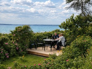 Een buitenruimte met eethoek en uitzicht op zee, omringd door weelderige begroeiing en bloeiende planten.