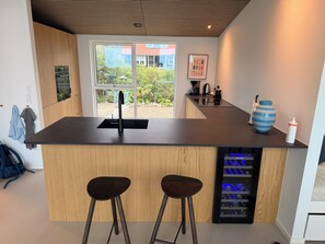 Eine moderne Küche mit einer zentralen Kochinsel, Barhocker, einem integrierten Weinkühler und Blick ins Freie.