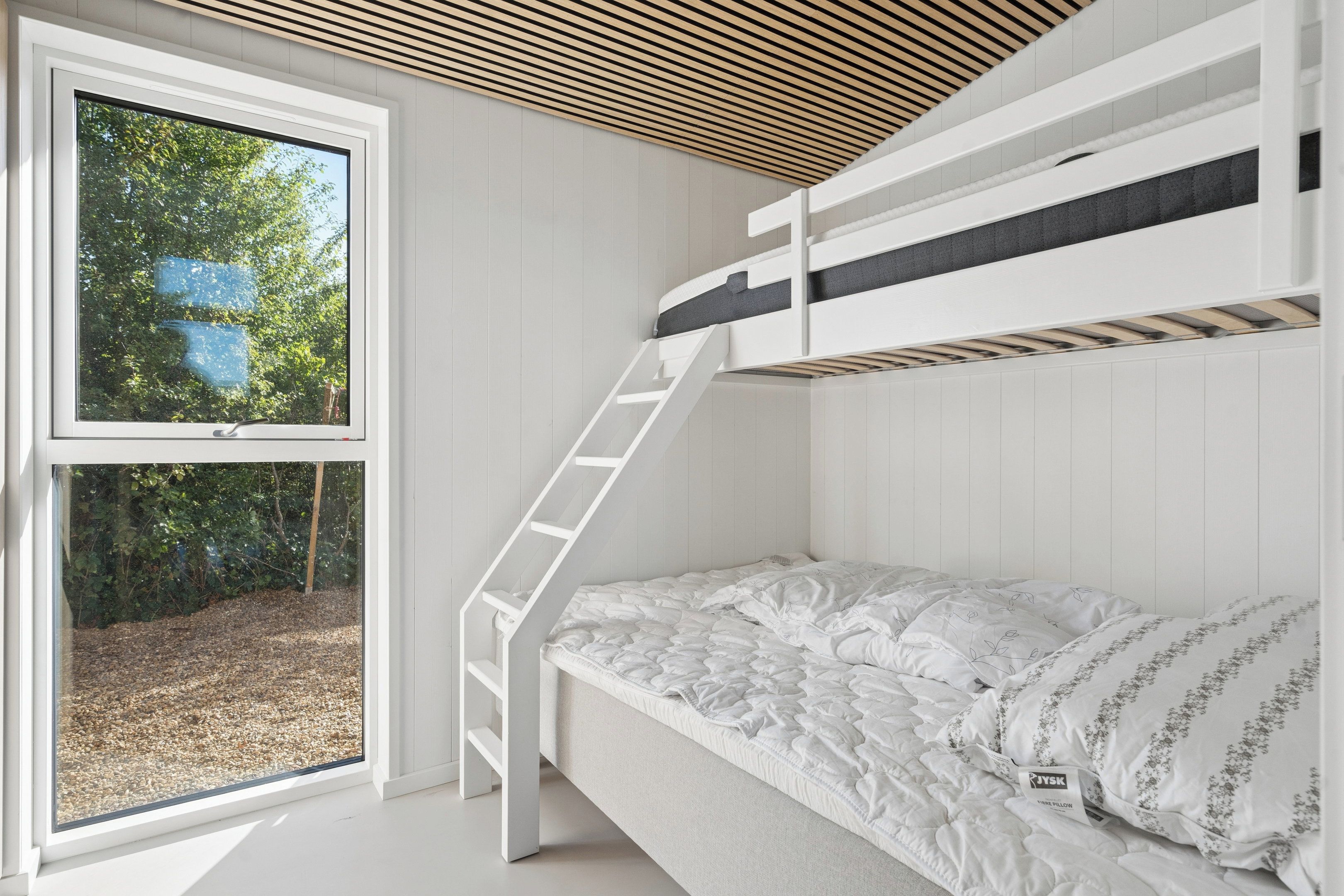 Un lit superposé avec une échelle, une fenêtre donnant sur les arbres et un mur blanc.