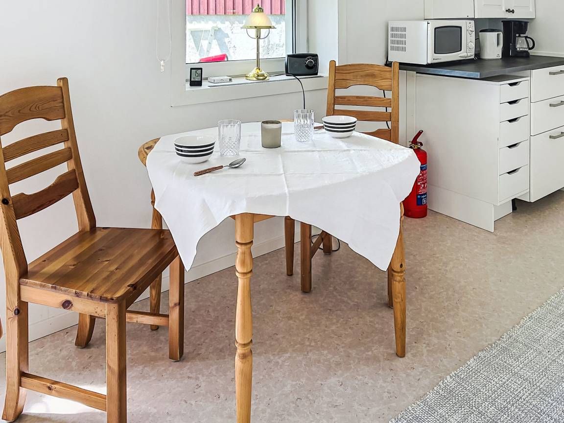 A dining area with a round table covered by a white tablecloth, wooden chairs, a small table lamp, and a microwave in the background.