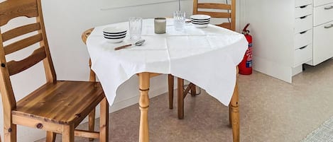 A dining area with a round table covered by a white tablecloth, wooden chairs, a small table lamp, and a microwave in the background.