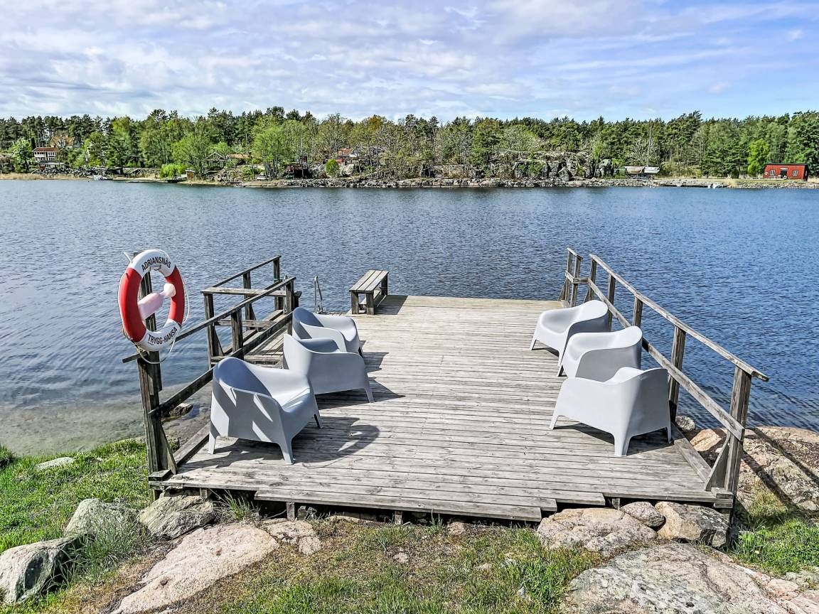 A wooden dock with white chairs and a lifebuoy, overlooking a calm lake with a forested shoreline.