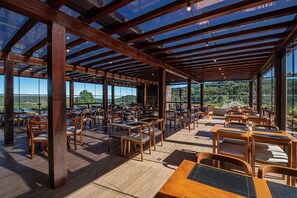 An open-plan restaurant with wooden tables and chairs, large windows offering a view of the landscape.