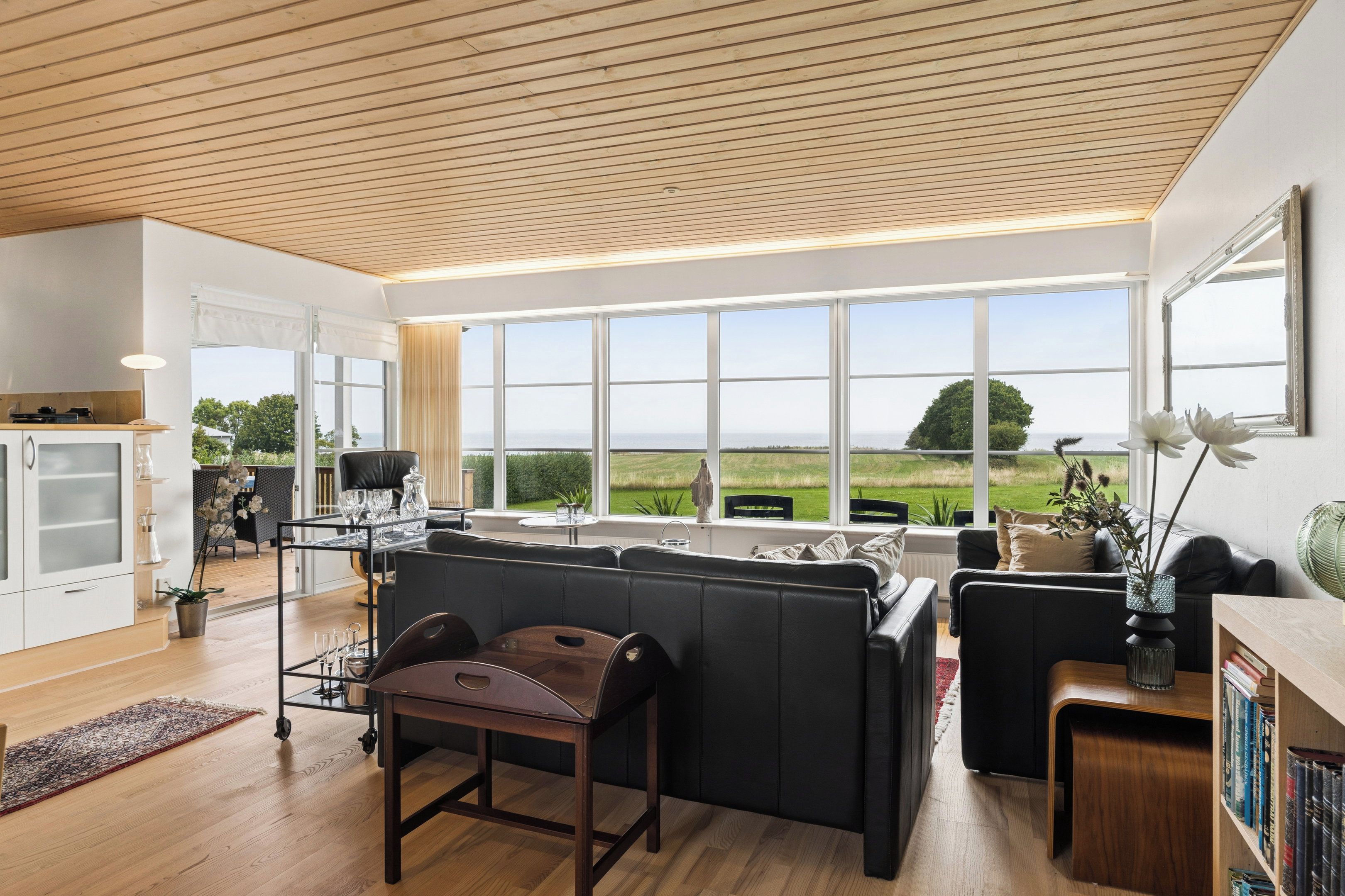 Een moderne woonkamer met een groot raam, een zwarte leren bank, een houten eettafel en een keuken met witte kasten.