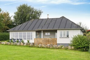 Yhden kerroksen talo, jossa on kalteva katto, etukuisti puukaitteella ja kivimuuri edessä.