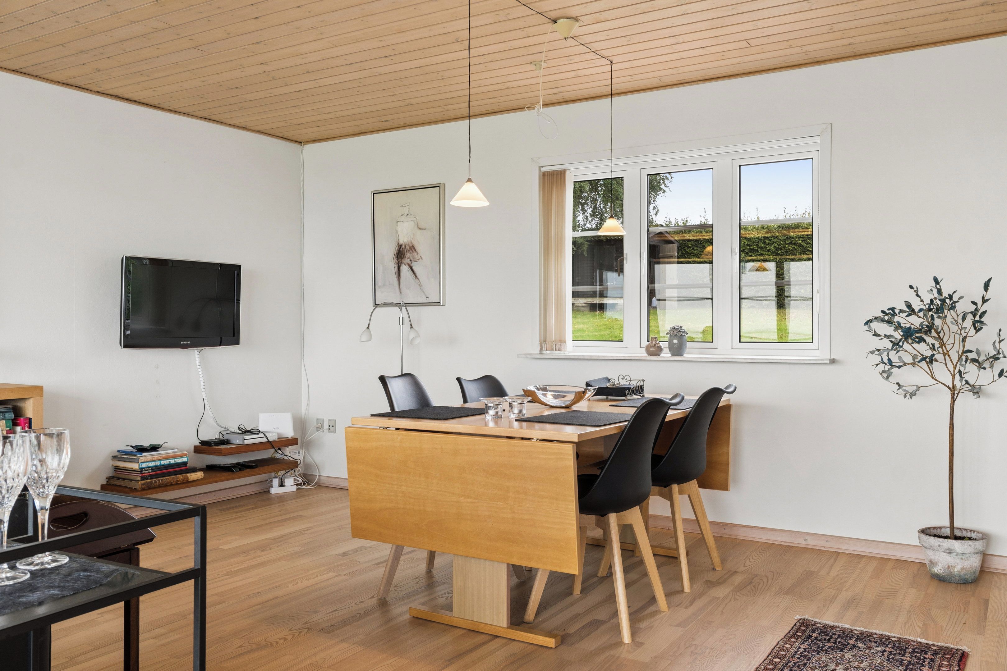 Un espace repas moderne avec une table en bois, des chaises noires, un téléviseur fixé au mur et une fenêtre donnant sur un paysage verdoyant.