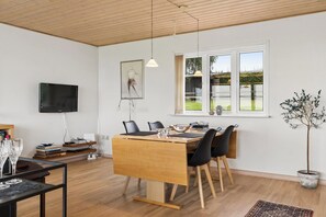 A modern dining area with a wooden table, black chairs, a television mounted on the wall, and a window with a view of greenery. - Holiday home near the water on beautiful Als (Augustenborg)
