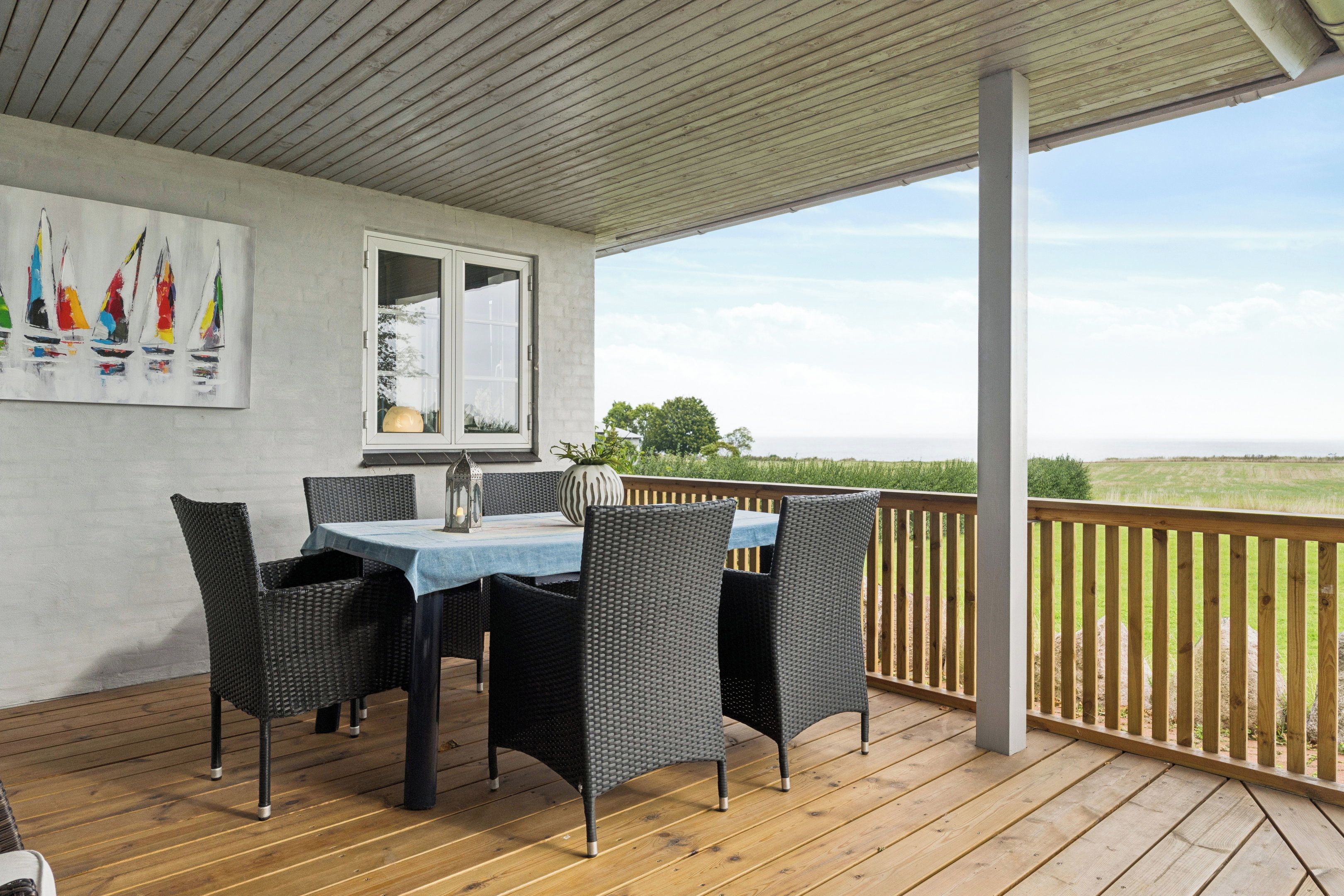 Een overdekt terras met een tafel en stoelen, uitzicht op het landschap, en een schilderij van zeilboten aan de muur.