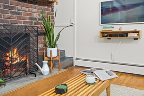 Een gezellige woonkamer met een open haard van baksteen, een houten salontafel, een kamerplant en een boekenplank.