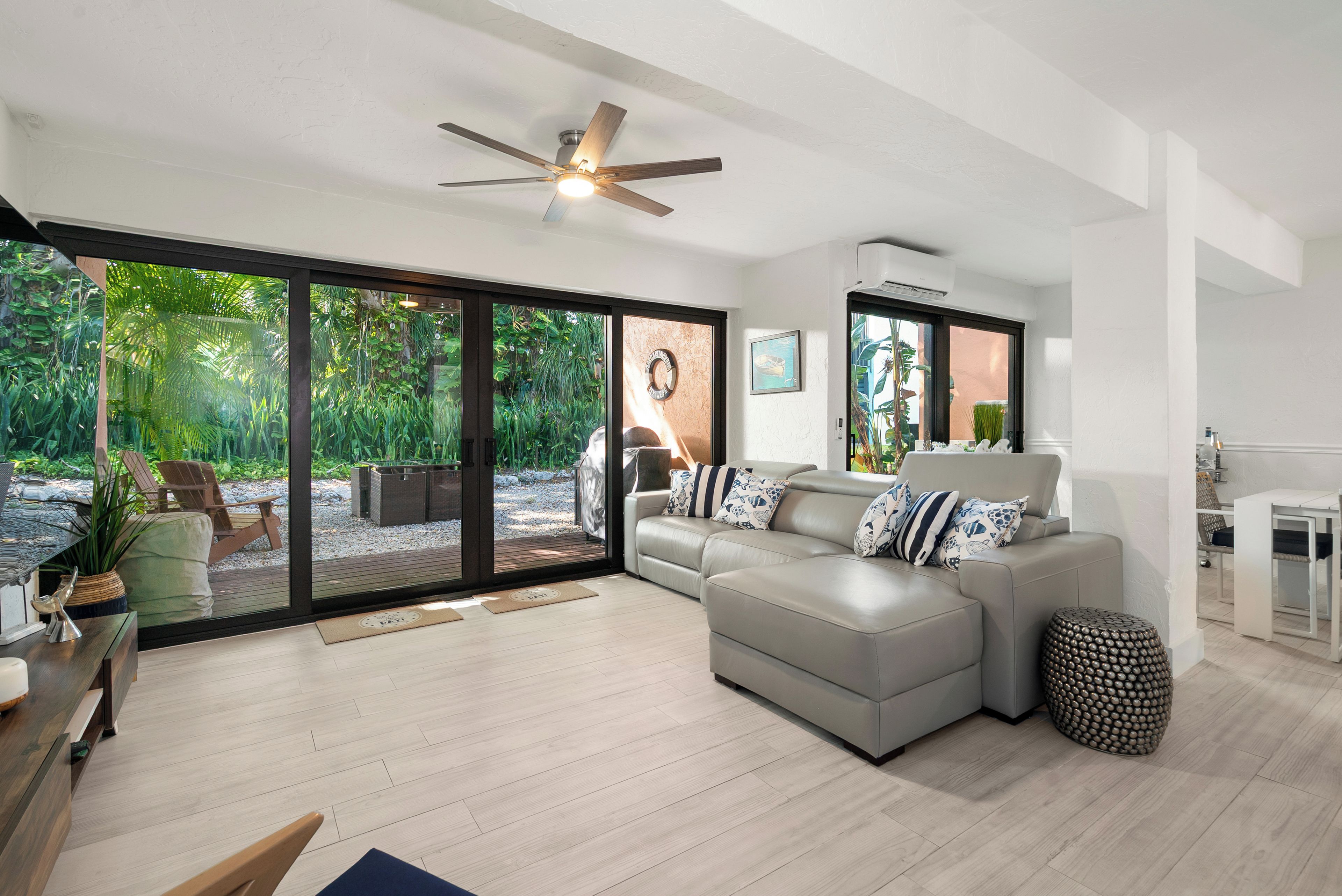 Un salon moderne avec un canapé d’angle gris, un ventilateur de plafond et une vue sur un jardin, visible  à travers de grandes portes vitrées.