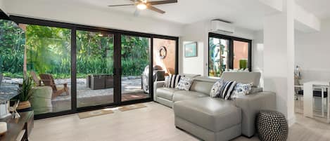 A modern living room with a grey sectional sofa, a ceiling fan, and a view of a garden through large glass doors.