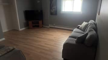 Una sala de estar con un sofá gris, piso de madera, un televisor sobre un mueble bajo, una ventana con persianas y un ventilador de techo.