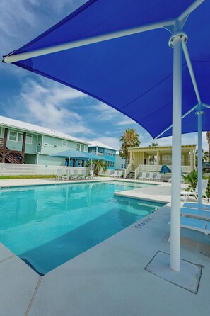 Piscina com espreguiçadeiras e guarda-sóis, cercada por edifícios com esquema de cores azul claro e branco.