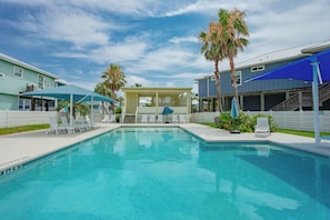 A swimming pool with lounge chairs and umbrellas, surrounded by palm trees and a covered seating area. - Shore Dream-Game Room/Community Pool/Mins to Beach (Port Aransas)