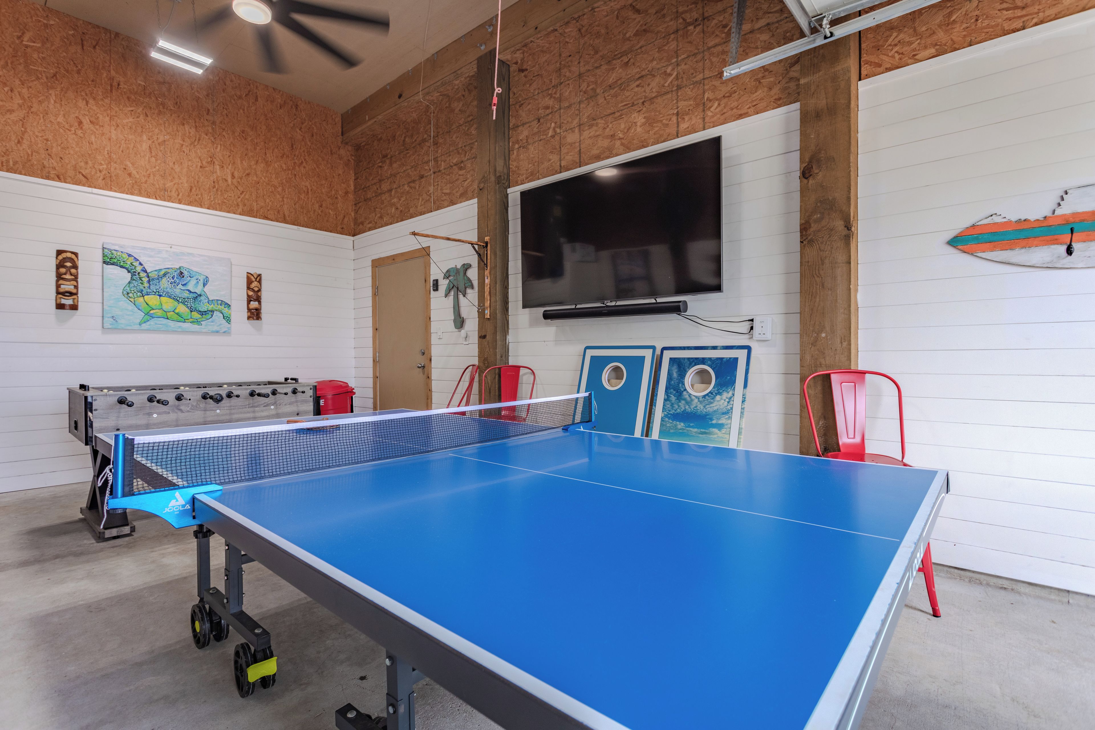 A ping pong table in a room with a wooden ceiling, a television mounted on the wall, and a surfboard decoration.