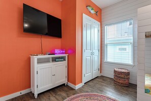 A room with a television mounted on the wall, a white cabinet with a neon sign, a door with a fish decoration, and a window with blinds. - Shore Dream-Game Room/Community Pool/Mins to Beach (Port Aransas)