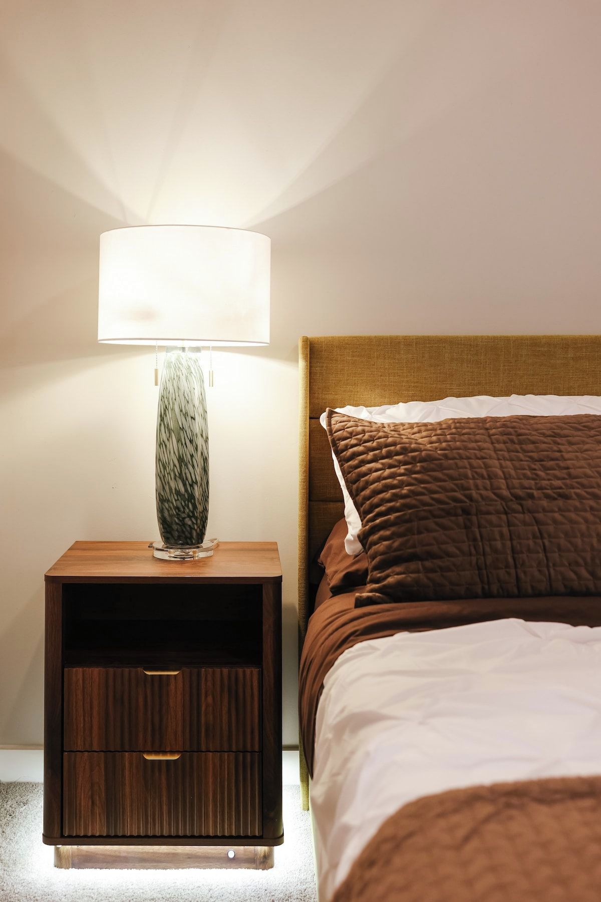 A neatly made bed with a brown quilt, a wooden nightstand with a lamp, and a drawer.