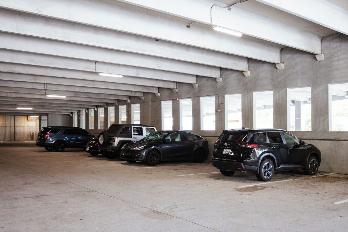 A parking garage with concrete pillars, overhead lighting, and large windows.
