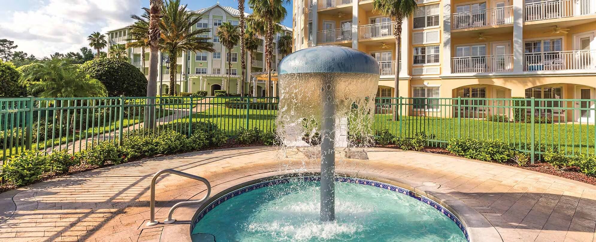 A swimming pool with a fountain, surrounded by a green fence, and a building with balconies in the background.