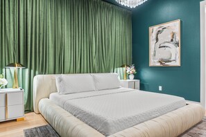 Een slaapkamer met een bed, groene gordijnen, een nachtkastje met een lamp en een schilderij aan de muur.