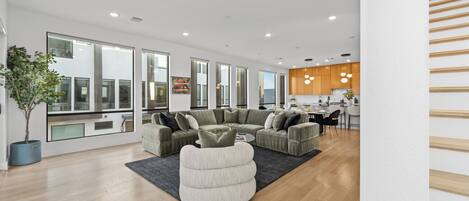 A modern living room with a sectional sofa, a round ottoman, and a large window offering a view of the city.