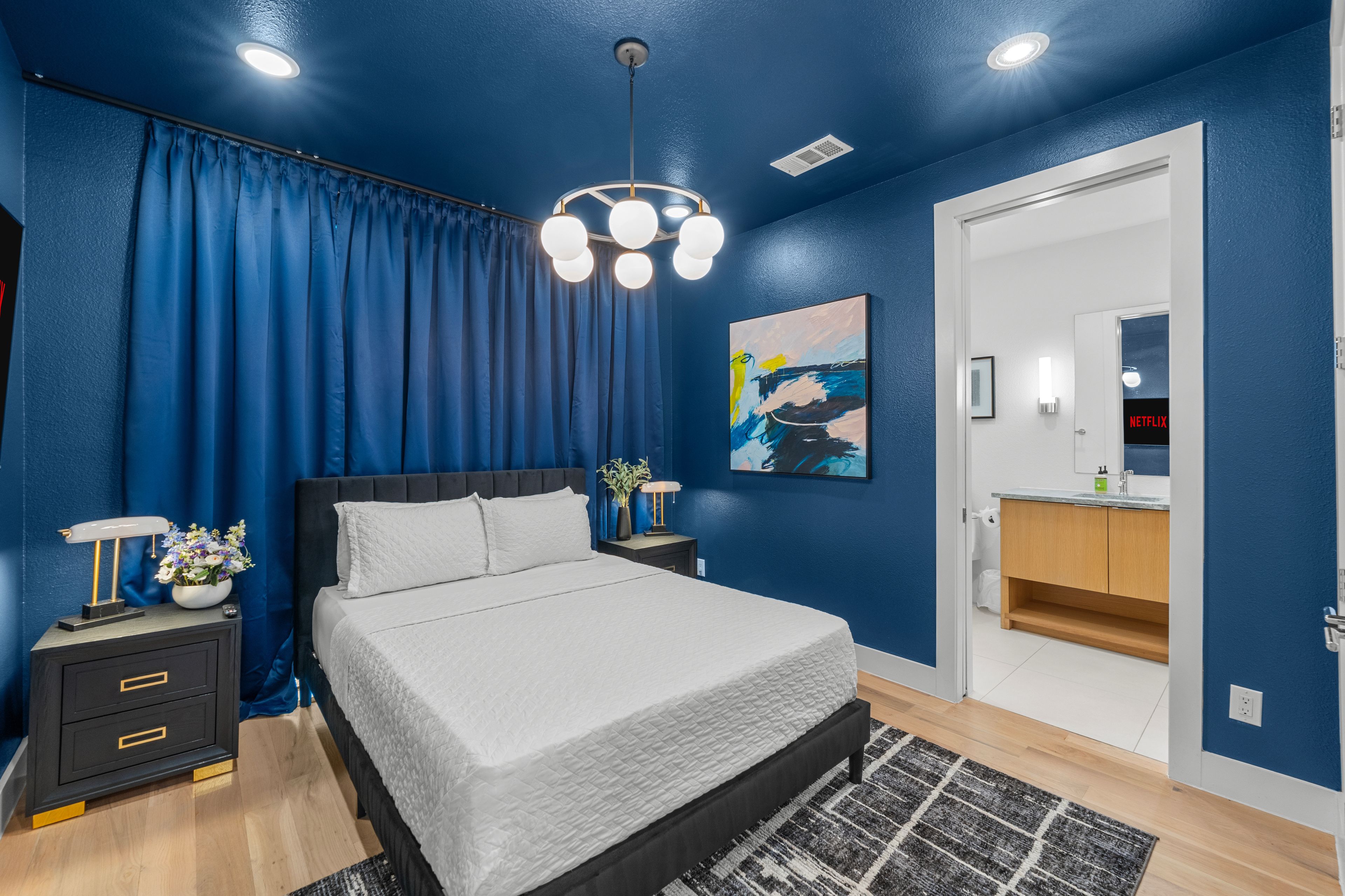 A bedroom with a bed, nightstands, a painting, a door, and a bathroom with a sink and mirror.