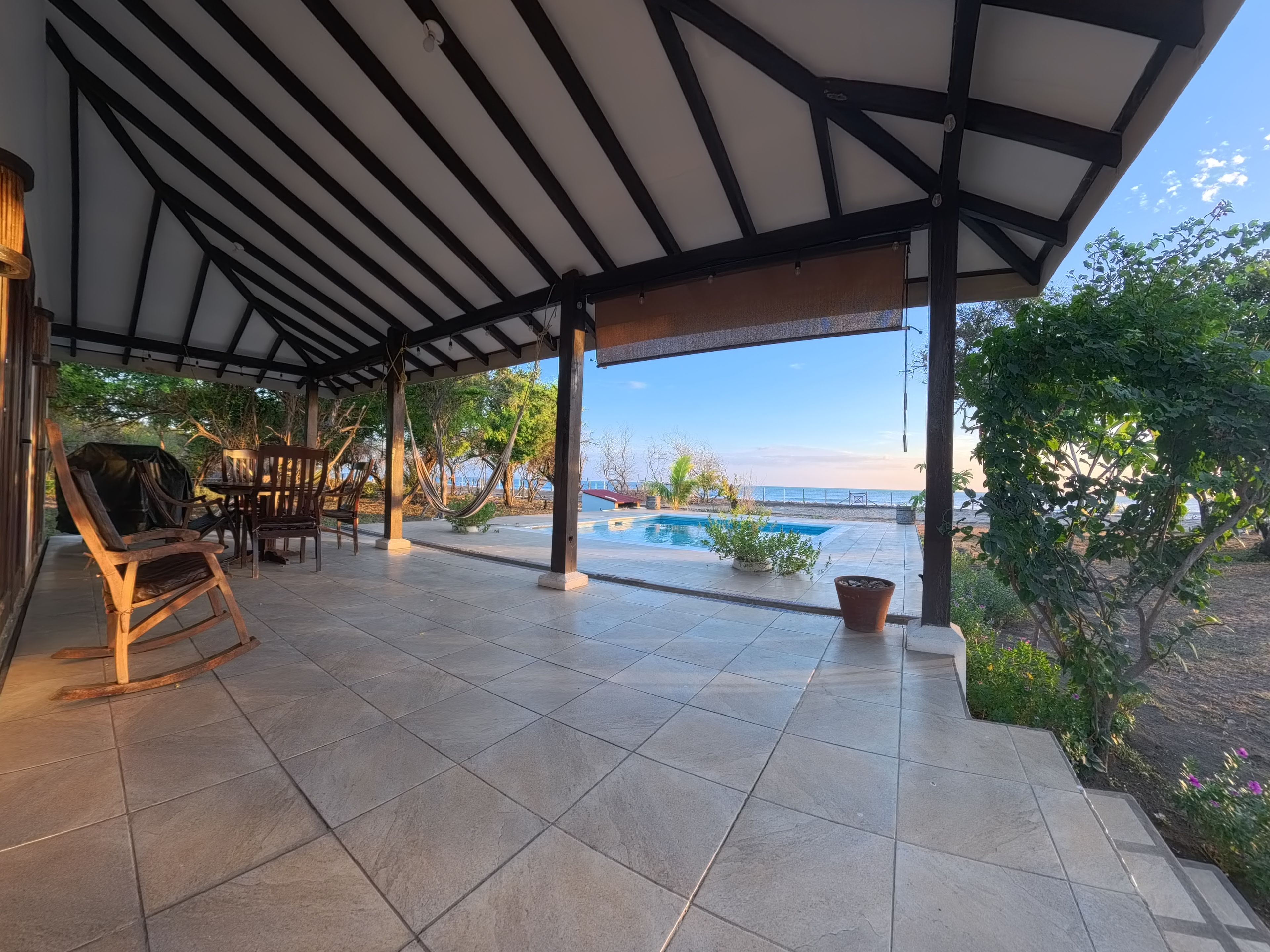 Un espacio exterior techado con piscina, mobiliario de madera y vistas al mar.