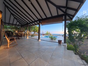 A covered outdoor area with a pool, wooden furniture, and a view of the sea.