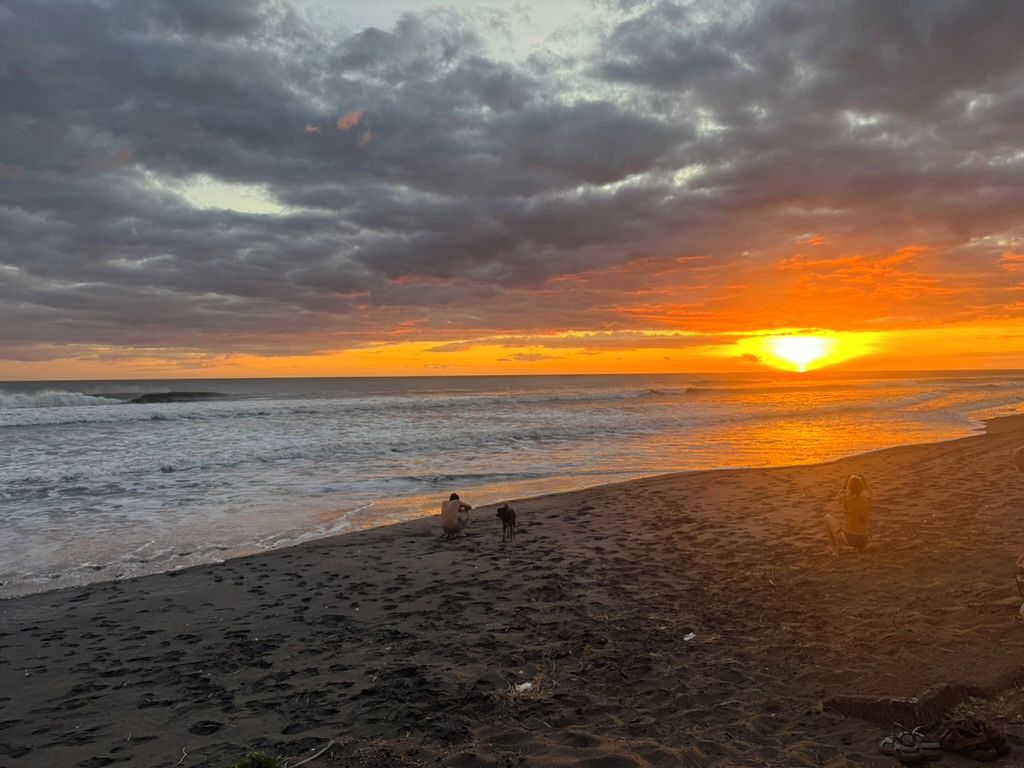 A beach with footprints leading to the water, a sunset in the background, and a cloudy sky.